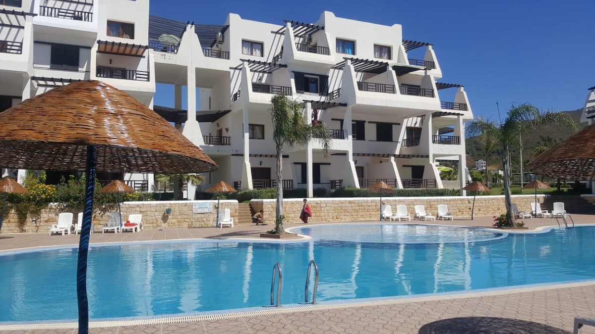 A spacious swimming pool is surrounded by lounge chairs and palm trees, set within a well-maintained garden area. The multi-story residence is visible in the background, featuring a clear blue sky above. Shaded areas are provided by thatched umbrellas near the pool.