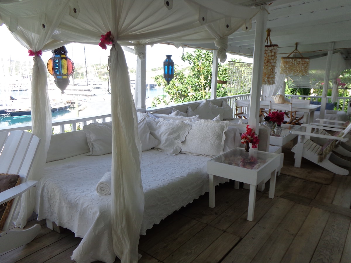 A spacious outdoor seating area is highlighted by soft white linens and cushions, creating a relaxed atmosphere. The area features white wooden furniture and decorative lanterns. Lush greenery surrounds the space, while the harbor view showcases boats docked nearby.