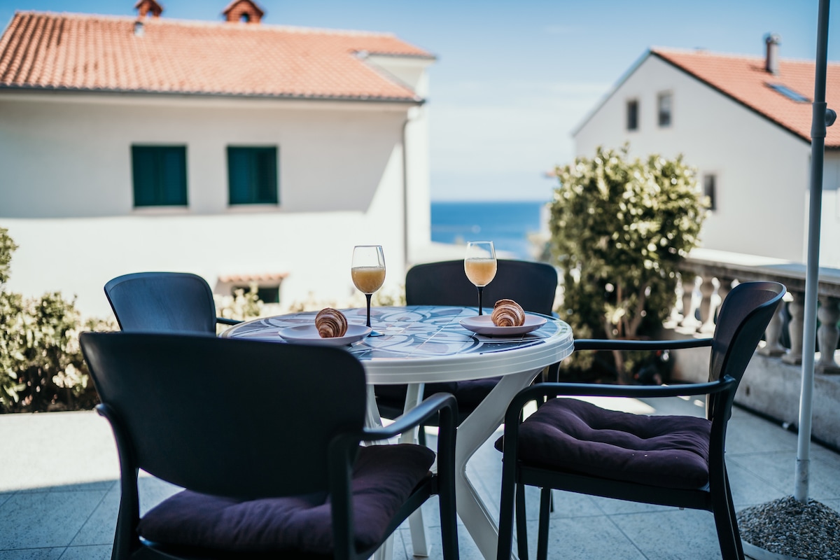 A terrace is showcased with a round table set for two, featuring two glasses of a light beverage and pastries. Black chairs with purple cushions surround the table, while a view of the sea and nearby houses is visible in the background.