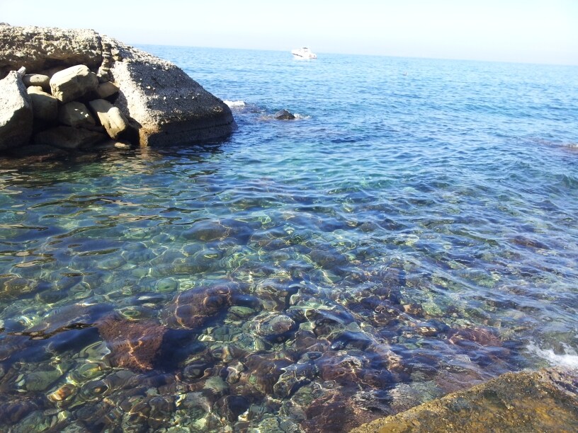 Clear turquoise waters gently lap against rocky shorelines, revealing a colorful underwater landscape. Sunlight dances on the surface, creating a shimmering effect above the stones and sea life beneath. A small boat can be seen in the distance, adding to the serene coastal atmosphere.