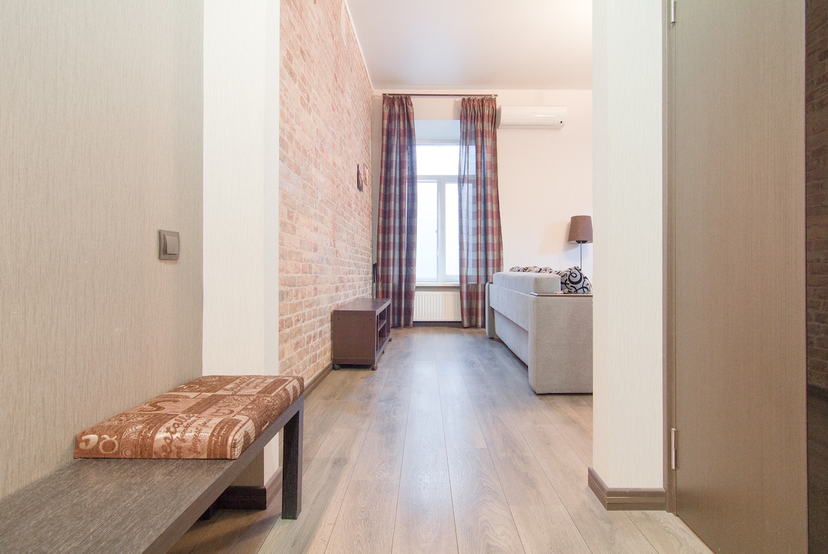 A hallway leads into a spacious living area featuring rustic brick walls. Natural light is let in through a large window, adorned with checked curtains. A comfortable gray sofa is positioned in the room, complemented by a small side table.