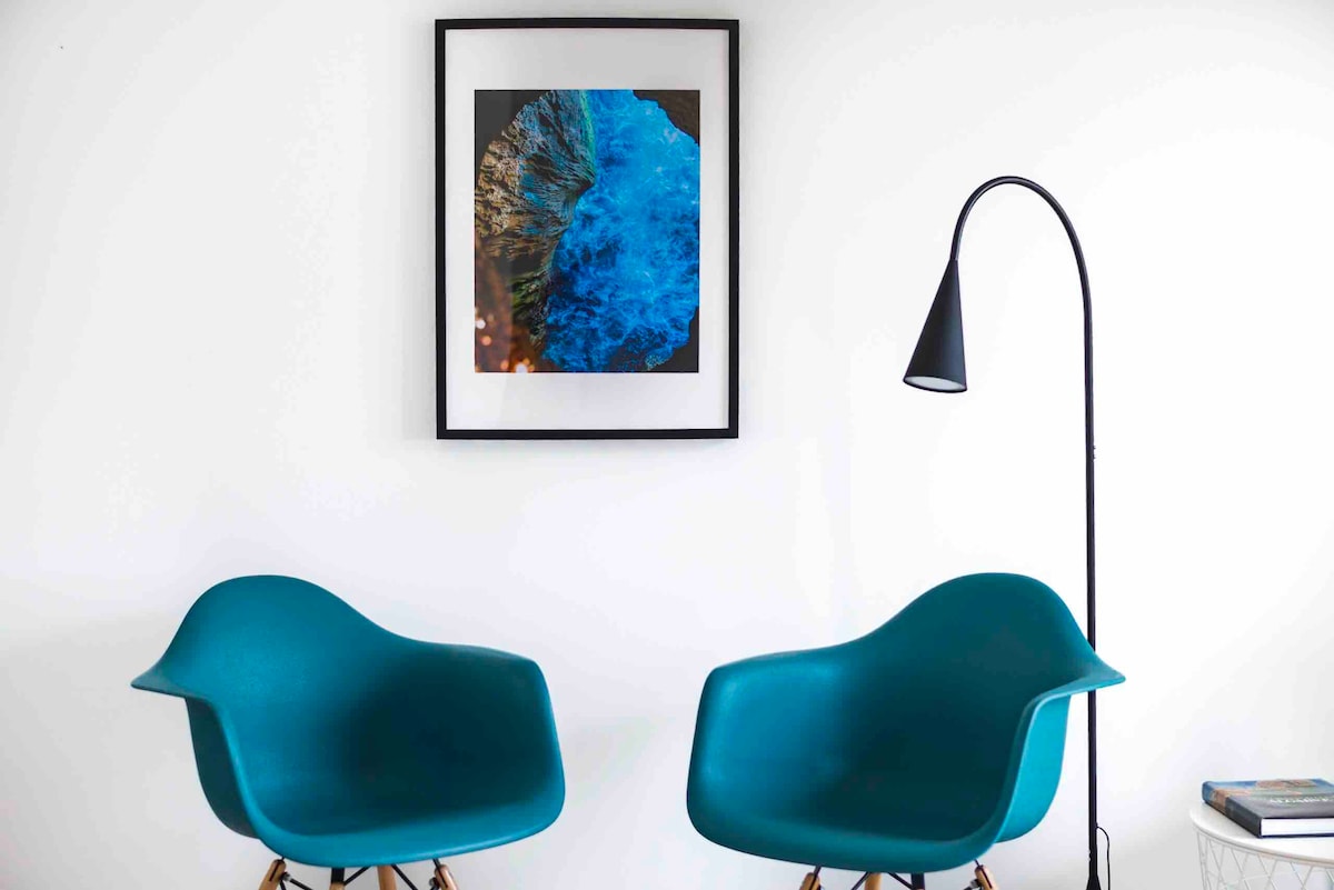 Two turquoise chairs are positioned beside a modern floor lamp. A framed artwork featuring a vibrant blue and brown design is displayed on a white wall, enhancing the minimalist decor of the space.