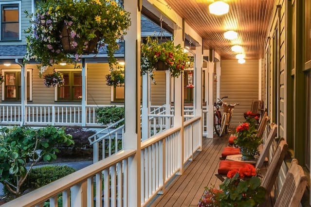 A welcoming covered porch features rocking chairs alongside vibrant flower pots. Hanging planters overflow with colorful blooms and gentle lighting creates a cozy ambiance. A bicycle is parked in the background, indicating accessibility to explore the surrounding area.