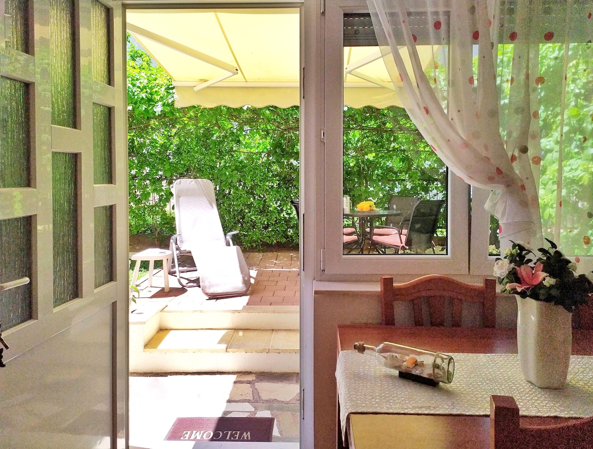 An open door reveals a private garden area, featuring a sun lounger and outdoor seating under a shaded canopy. A table set up for dining is visible, complemented by a vase of flowers. Lush greenery provides privacy and a serene environment.