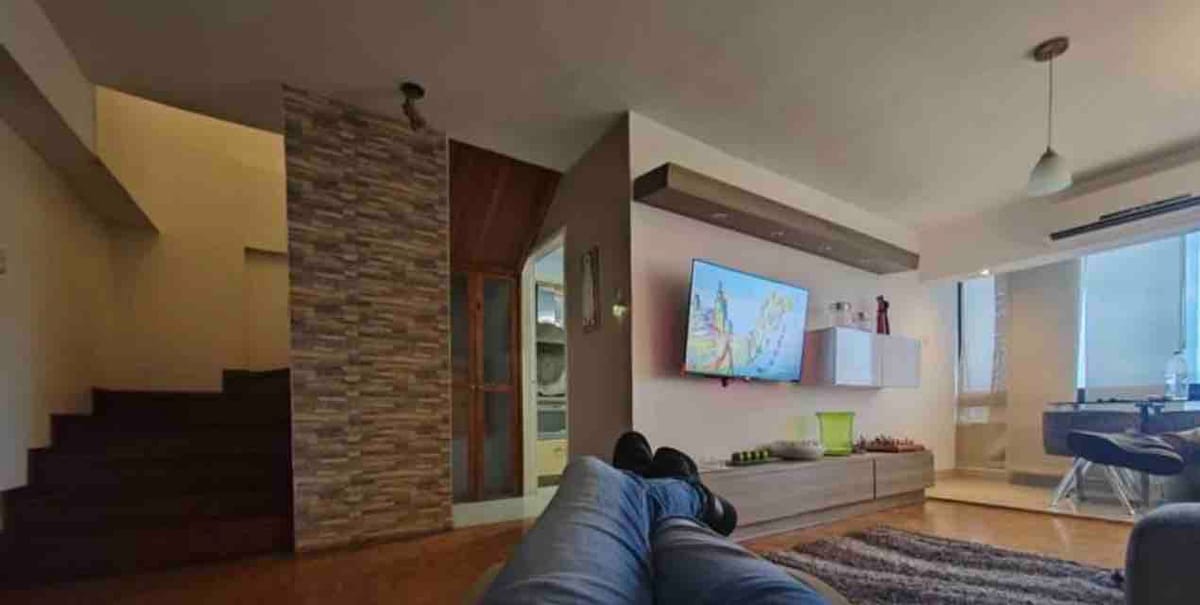 A cozy living area is featured, displaying a mounted flat-screen TV on a light-colored wall unit. Natural light enters through large windows, illuminating the wooden floor. A staircase leads to the upper level, while a glimpse of the kitchen is visible in the background.
