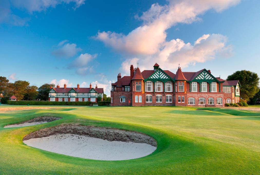 A traditional golf club building with distinctive green and white timber features is set against a blue sky. Well-maintained grass and sand traps surround the structure, creating an inviting landscape for golf enthusiasts.