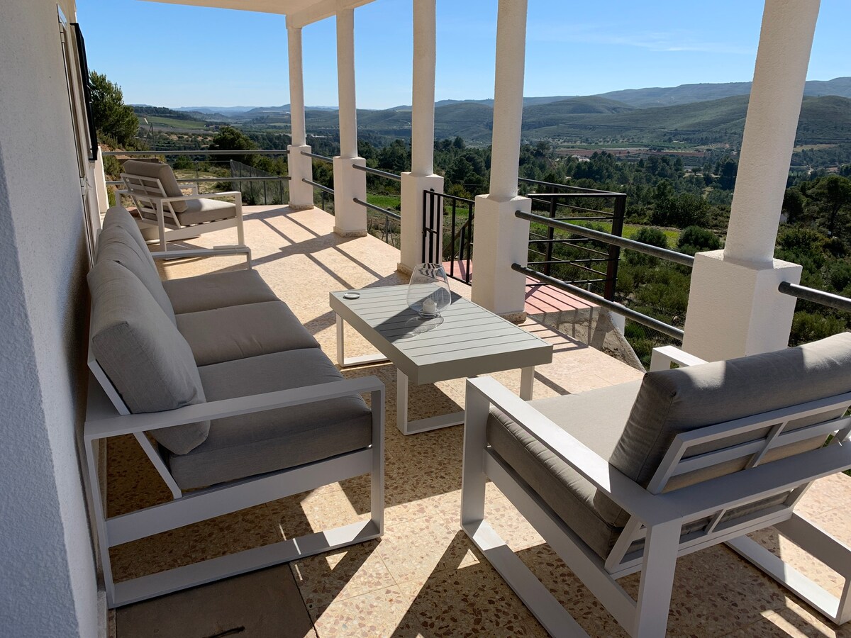 A spacious terrace is furnished with light-colored seating and a coffee table. The area offers a panoramic view of the surrounding hills and valleys, with sunlight casting soft shadows on the surface. The structure is supported by tall columns, providing a modern architectural touch.