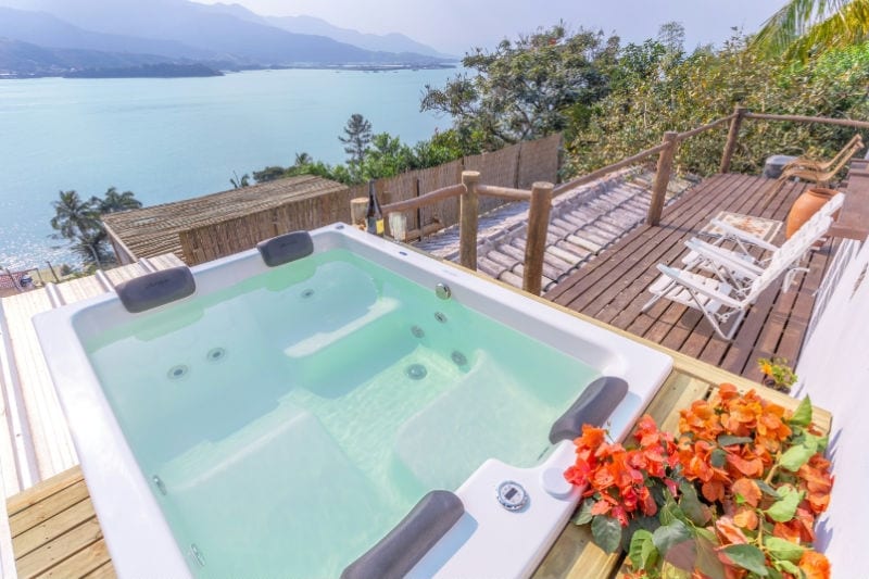 A private outdoor hot tub is positioned on a wooden deck, overlooking a scenic bay. Lush greenery and vibrant flowers surround the area, while distant mountains create a serene backdrop. Two white lounge chairs provide additional seating options, enhancing the tranquil environment.