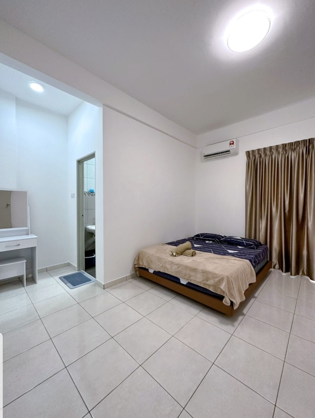 A bright sleeping area features a spacious bed covered with a neutral blanket. A small dressing table is positioned nearby, and a bathroom door is visible in the background. Floor tiles are light-colored, and soft curtains frame a window, allowing natural light to enter the room.