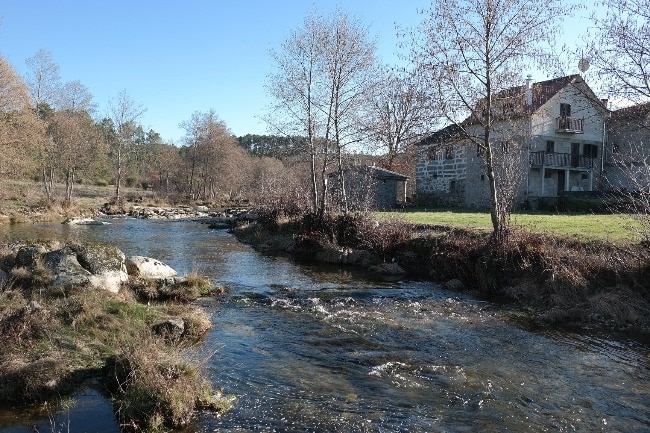The image depicts a serene riverside scene, with a gently flowing river bordered by smooth stones and grassy banks. A two-story stone house stands nearby, partially obscured by blossoming trees, under a clear blue sky. The peaceful landscape invites outdoor exploration.