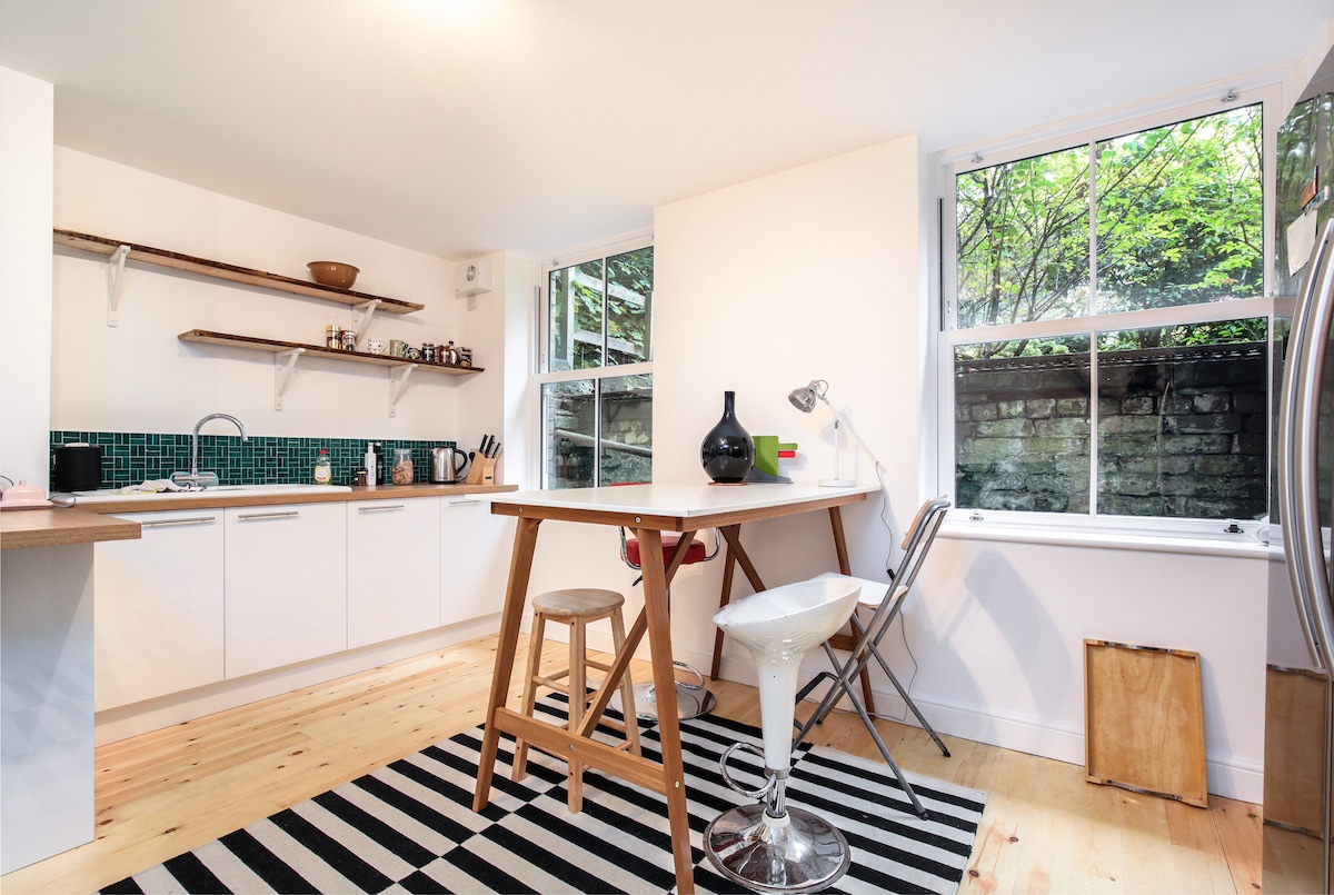 A bright kitchen area is showcased with modern cabinetry and a tiled back wall. A small table with stools occupies the center, beside a large window that invites natural light. A striped rug adds contrast to the wooden floor, while potted greenery is visible outside.