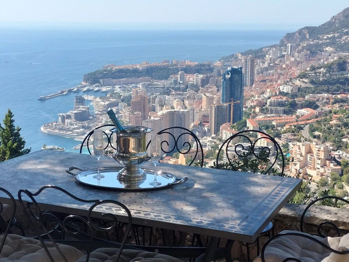 A dining table with a polished metal surface is set atop a stone terrace, showcasing a panoramic view of Monaco and the Mediterranean Sea. An elegant champagne bucket is positioned on the table, enhancing the scenic backdrop of the coastal city and nearby hills.