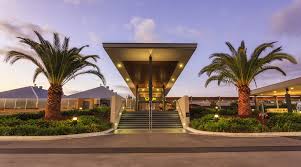 The entrance of a contemporary building is showcased with a large overhang supported by columns. Palm trees frame the approach, and well-manicured landscaping lines the pathway leading to the stairs. Warm lighting illuminates the facade, creating a welcoming ambiance during twilight.