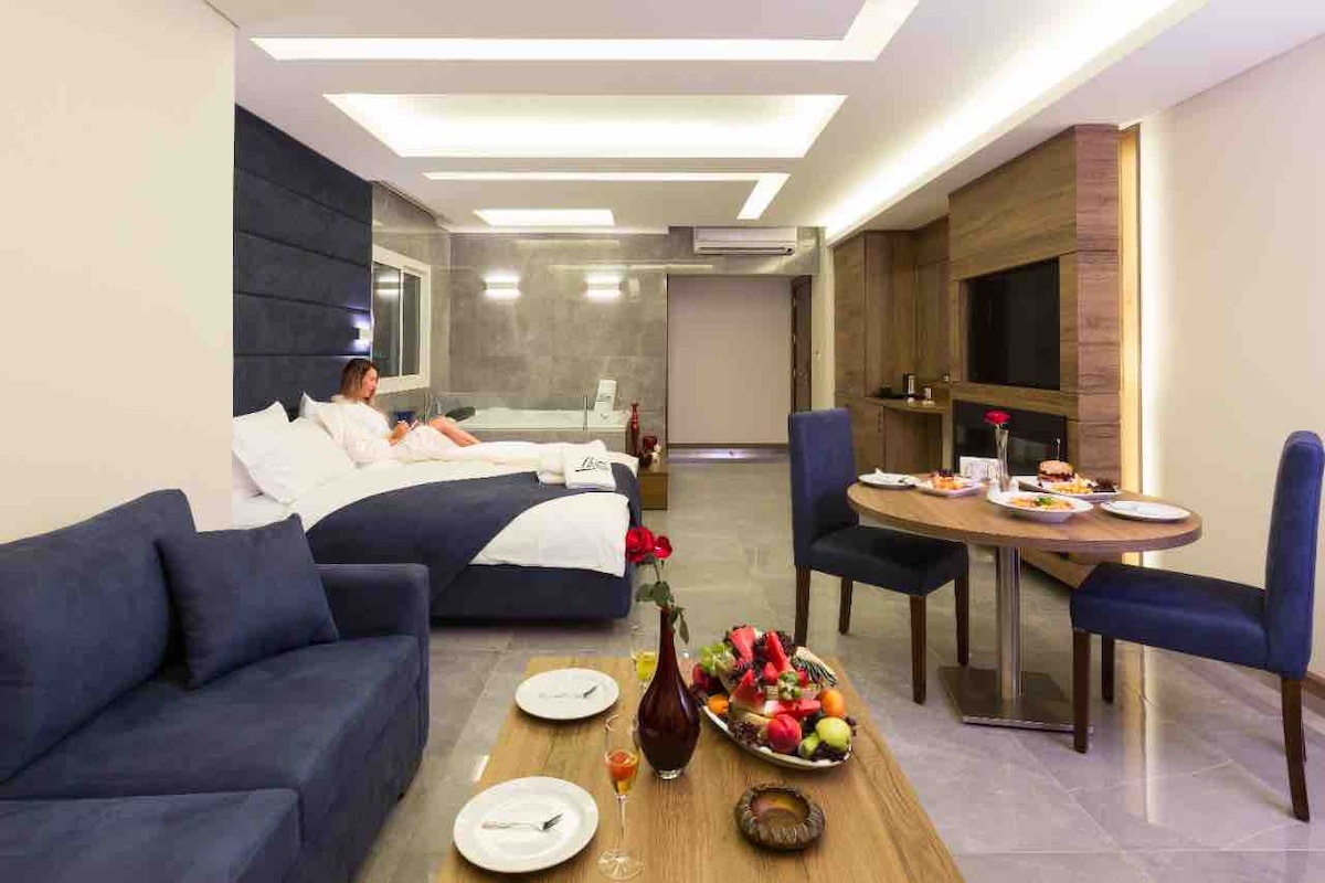 A modern living space features a comfortable sofa, a dining set with two chairs, and a table adorned with fresh fruit. A spacious bed is visible in the background, complemented by soft lighting and neutral tones throughout the room.