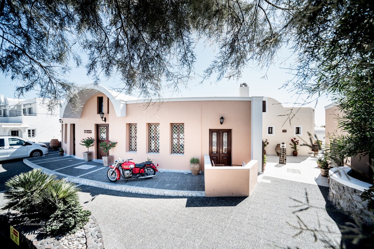 A charming exterior of a family-run accommodation is displayed, featuring a smooth stone pathway leading to an inviting entrance. Potted plants are placed by the windows, and a vintage motorcycle sits beside the entrance, adding character to the overall facade.