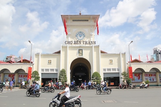 The bustling Ben Thanh Market is captured in the image, featuring an iconic clock tower flanked by vibrant red flags. Motorbikes are seen passing by, illustrating the dynamic atmosphere of the surrounding area. The structure showcases a blend of historical architecture and contemporary urban life.