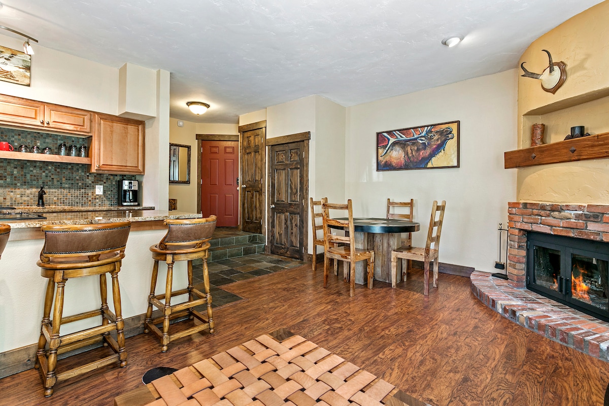 An open living area features a circular dining table with wooden chairs beside a cozy fireplace, set against a brick wall. The kitchen has a bar with stools, and a rich wooden door leads to an entryway. Natural light accentuates the warm tones of the space.