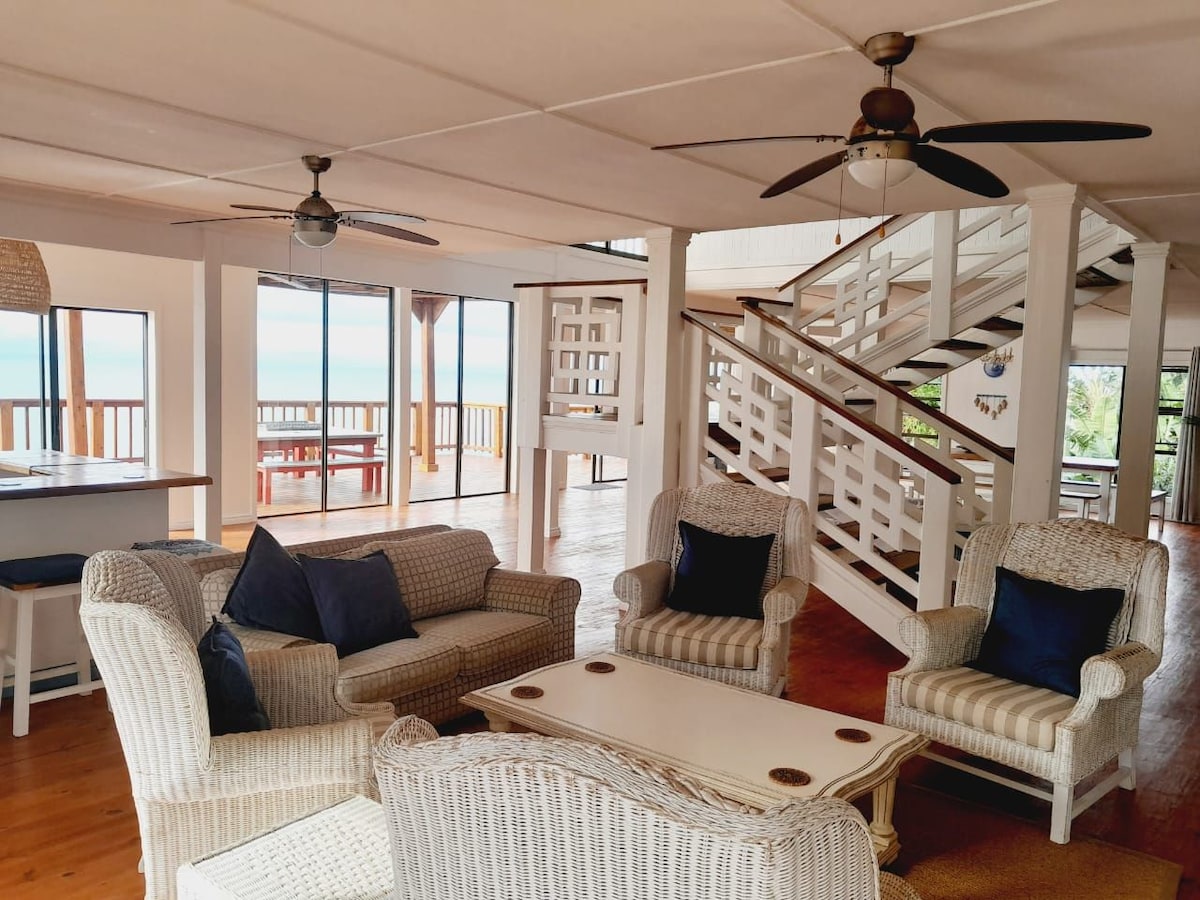 An open living area features comfortable seating arranged around a central table. Ceiling fans provide a cooling effect, while large windows showcase expansive views of the outdoors, inviting natural light into the space. A staircase leads to an upper level.