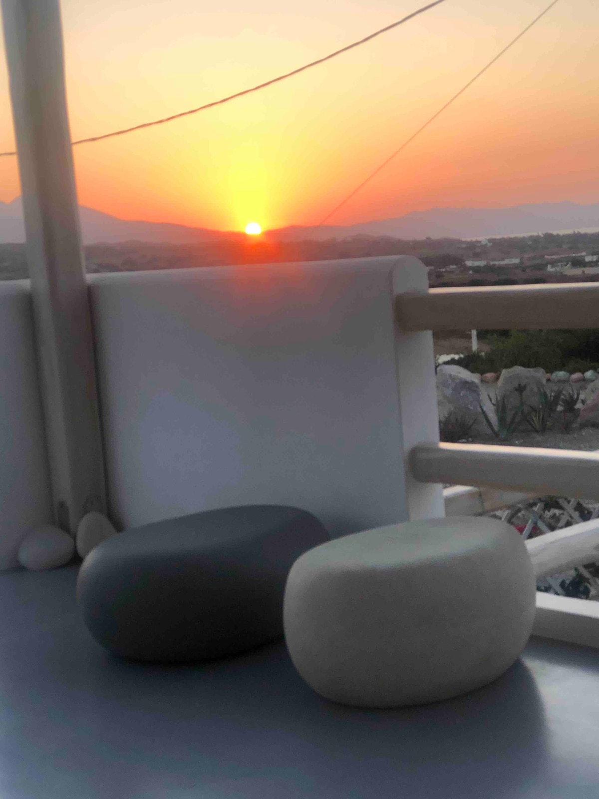 A balcony lounge area is captured with two round stools in neutral tones, facing a breathtaking sunset. The sun sets behind distant mountains, casting warm hues across the sky, while the serene landscape is framed by the balcony's railing.