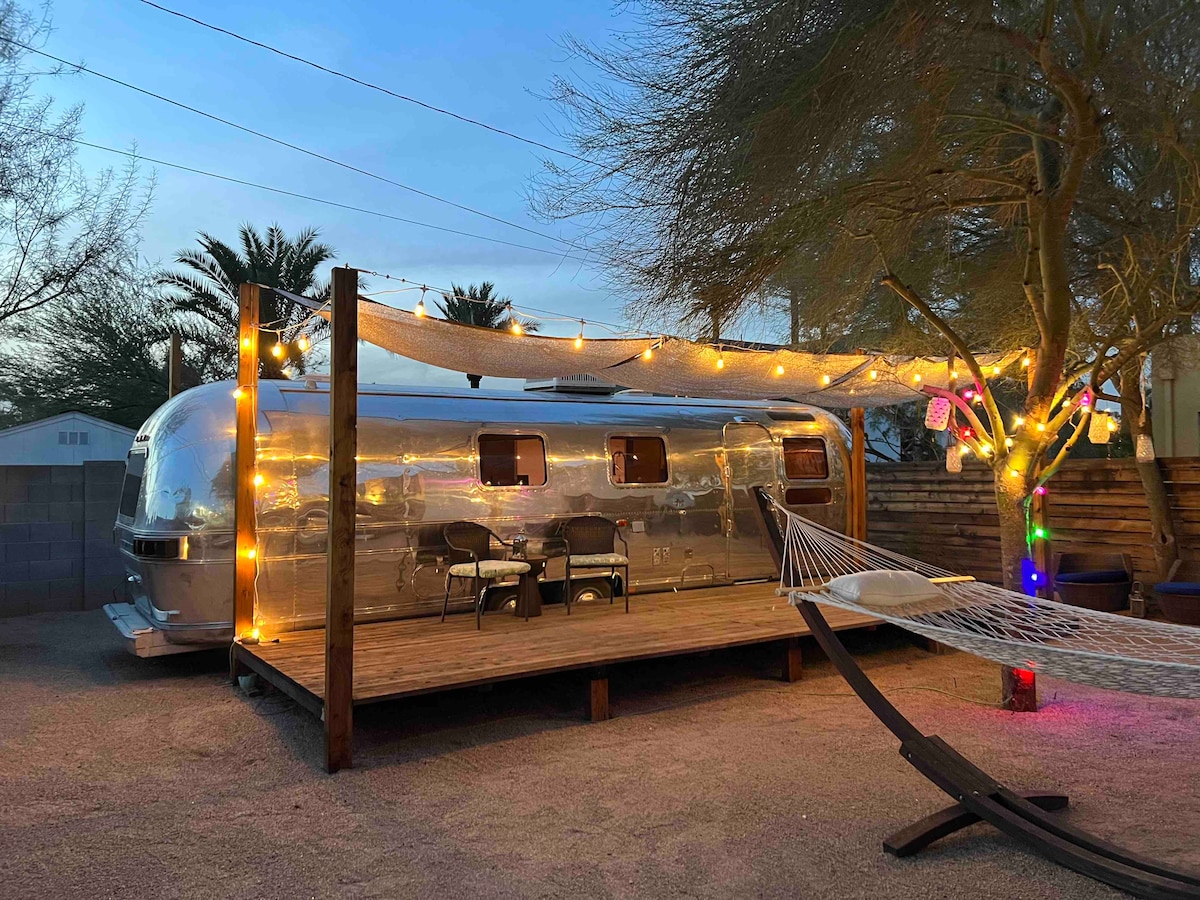An outdoor deck surrounds a vintage Airstream, illuminated by string lights. Two chairs are placed on the deck under a shaded canopy. A hammock is positioned nearby, enhancing the relaxed atmosphere of the space, with palm trees in the background.