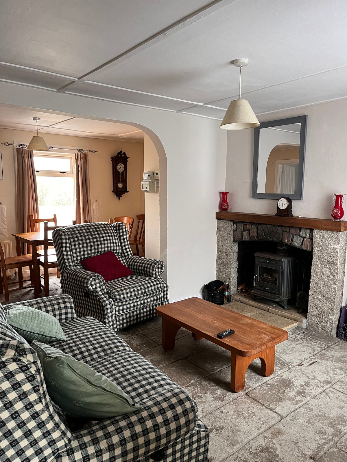 A living area features a checkered armchair and a matching sofa, complemented by a wooden coffee table. A small fireplace is set into the stone wall, and a mirror hangs above it. Natural light enters through a window with light curtains, illuminating the space.