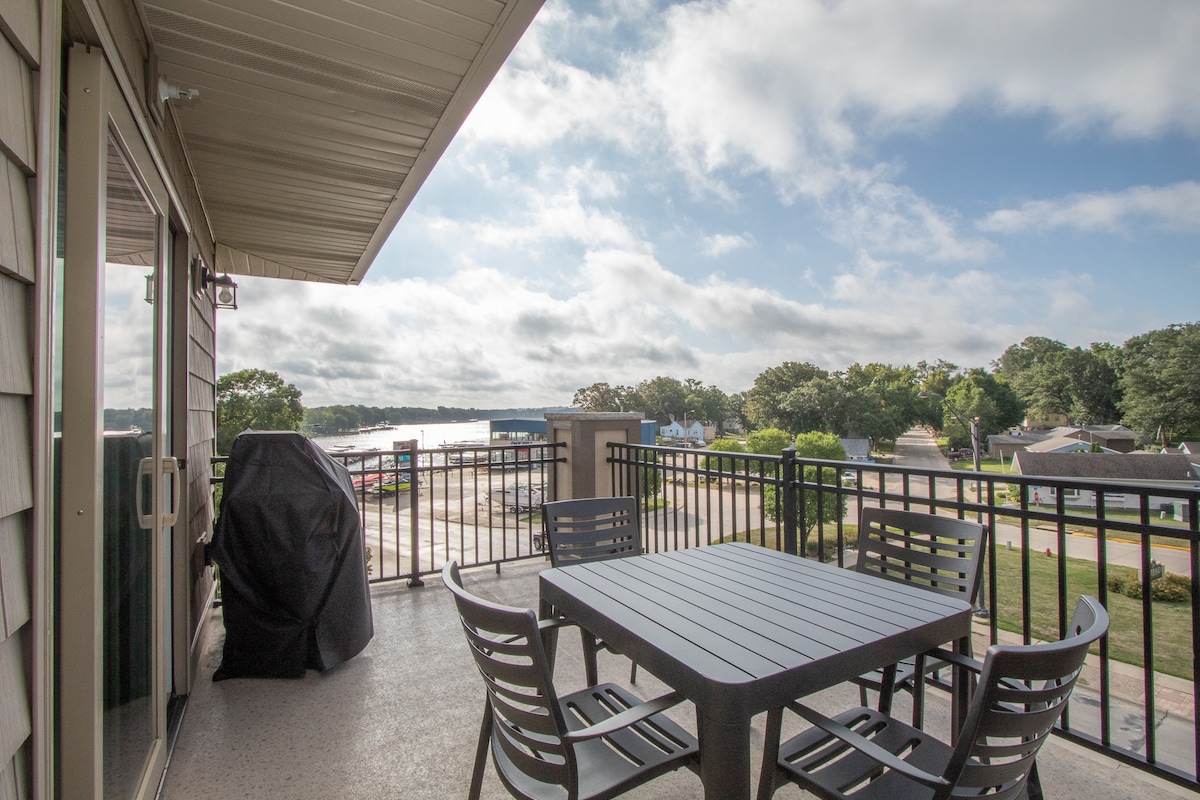An outdoor patio is presented with a dining table and several chairs arranged for gatherings. A grill is positioned nearby, and views of Lake Okoboji's boat launch and surrounding greenery are visible. The sky features soft clouds and natural light enhances the space.