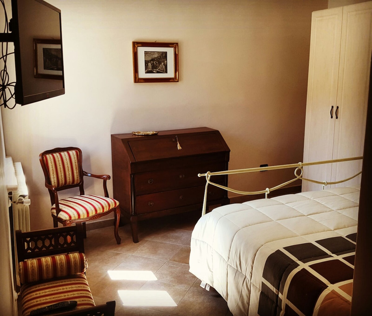 A tranquil sleeping area features a spacious bed draped in light bedding. A vintage wooden dresser stands opposite the bed, accompanied by a striped chair. A wall-mounted television adds convenience, while soft natural light enters through a nearby window.