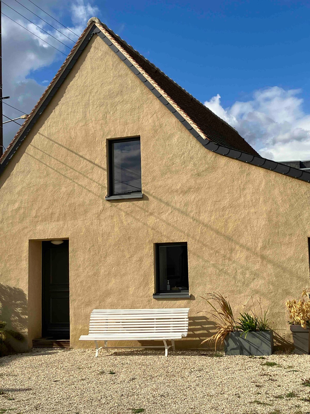 A quaint exterior view of a charming house displays a textured beige wall and a steeply pitched roof. A white bench is positioned on the gravel in front, complemented by two planters containing dried plants. Large windows provide natural light to the interior.