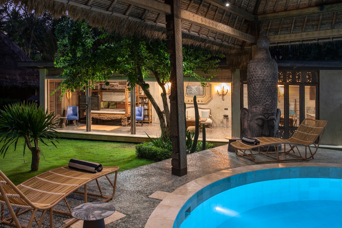 A private swimming pool is surrounded by a stone deck with wooden lounge chairs. The open-plan living space is visible through expansive glass doors, revealing a cozy bedroom and stylish furnishings. Lush greenery and a large carved statue enhance the tranquil atmosphere.