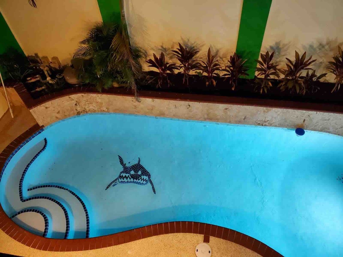 The pool area is outlined with elegant tile work, featuring a unique design of a fish at the bottom. Surrounded by lush greenery, the clear blue water is inviting, with a spacious hot tub seamlessly integrated into the design.