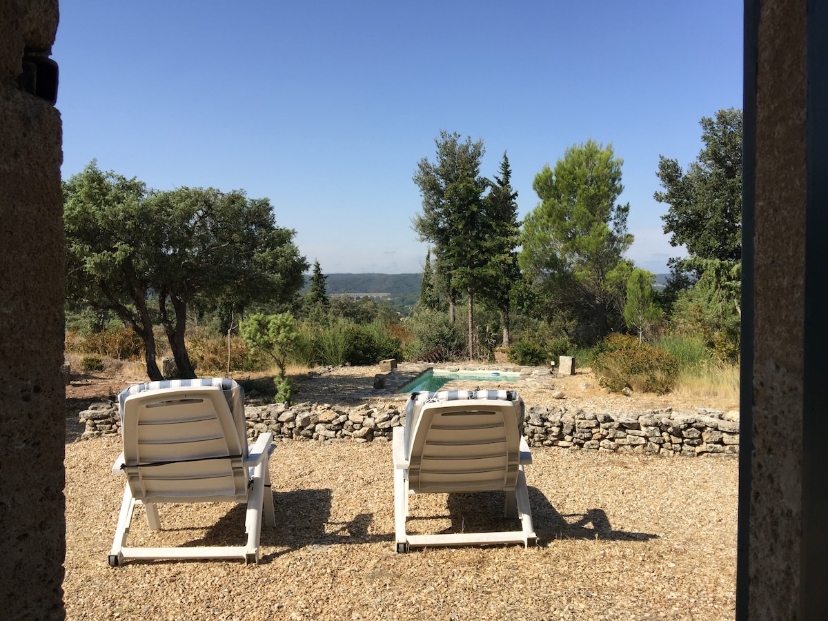 Two lounge chairs are positioned on a gravel patio, facing a serene view of rolling hills and greenery. Beyond the seating area, a small swimming pool reflects the clear blue sky, surrounded by Mediterranean vegetation.