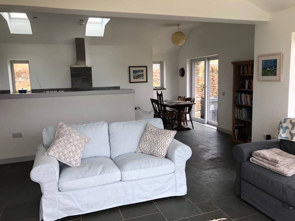 An open-plan living area features a light blue couch with decorative pillows, slate flooring, and a modern kitchen in the background. A dining set is positioned nearby, with bookshelves filled with literature and a large window allowing natural light into the space.