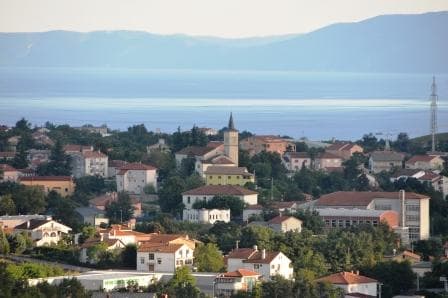 A scenic view of the Marinici area is presented, showcasing a mix of residential buildings and greenery. The distant horizon features the blue expanse of the sea, while a church spire is visible among the rooftops, contributing to the tranquil ambiance of the surroundings.