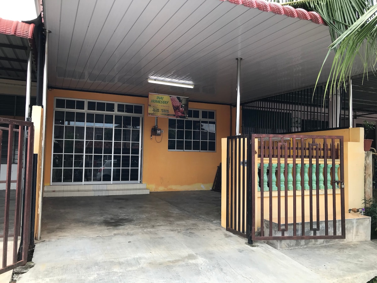 The image displays a covered carport with a concrete floor, featuring a prominently adorned orange wall. Large glass windows are visible, providing natural light to the interior space. A gated entrance is present, flanked by decorative green railings.