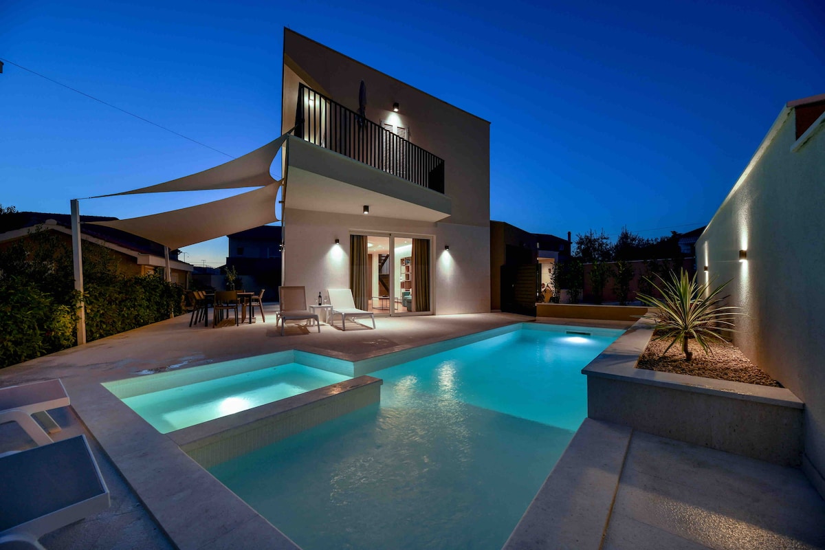 An outdoor area is showcased with a modern pool illuminated in soft blue hues under the evening sky. A hydromassage section is visible, along with a shallow area for children. Comfortable deck chairs are positioned nearby, adding to the inviting atmosphere of the space.