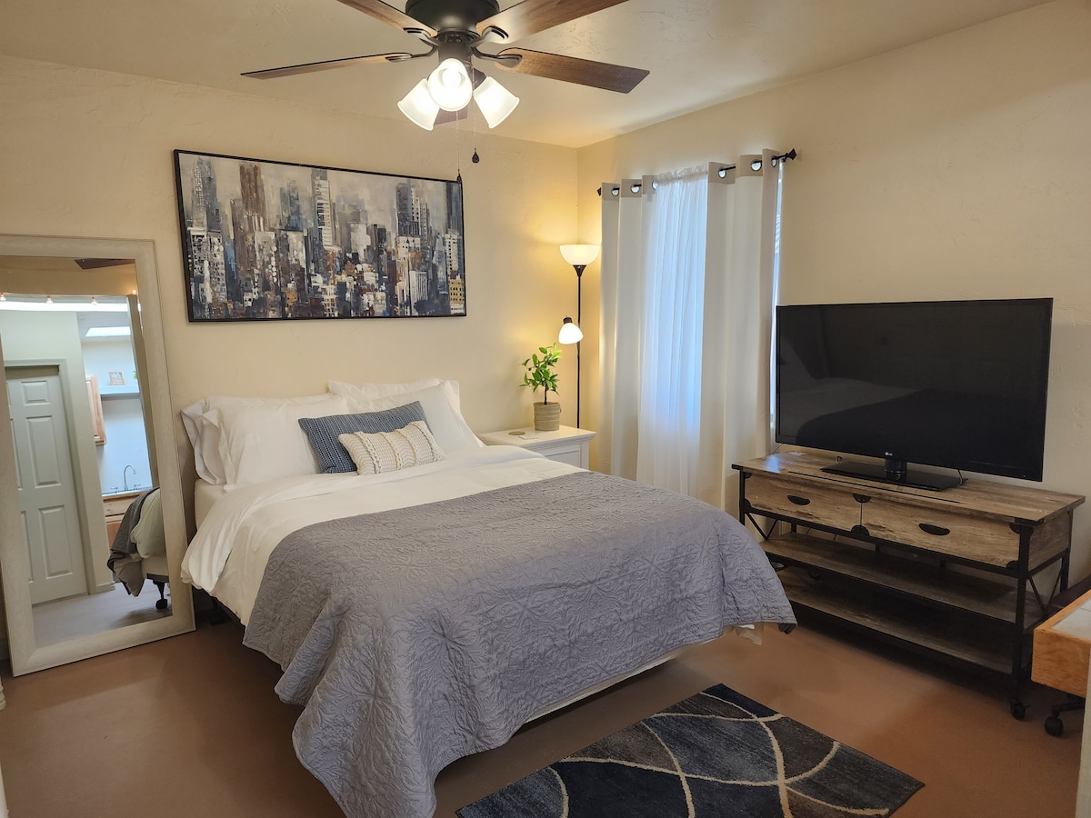 A spacious sleeping area features a large bed with a light quilt cover. Accompanying furniture includes a wooden TV stand with a television, and a bedside lamp casts soft light. Natural light filters through a window adorned with sheer curtains, complemented by wall art of a cityscape.