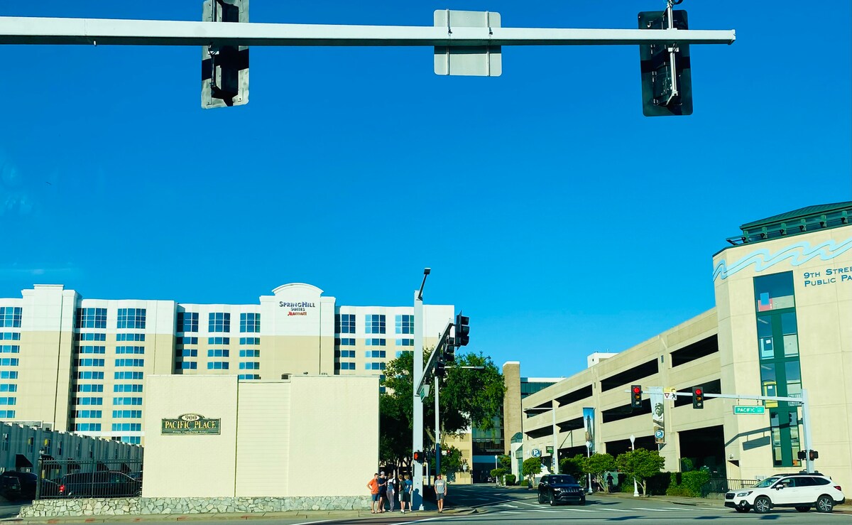 A busy intersection is captured with a clear blue sky above. Pedestrians are seen waiting at a traffic light, with various buildings visible in the background, including a hotel and a parking garage, indicating a vibrant urban area. Traffic signals are prominently displayed.