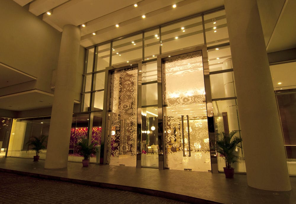 The entrance features large glass doors that reveal a modern lobby with intricate, shimmering wall decor. Soft lighting casts a warm glow, creating an inviting atmosphere. Potted plants flank the entrance, enhancing the contemporary design.
