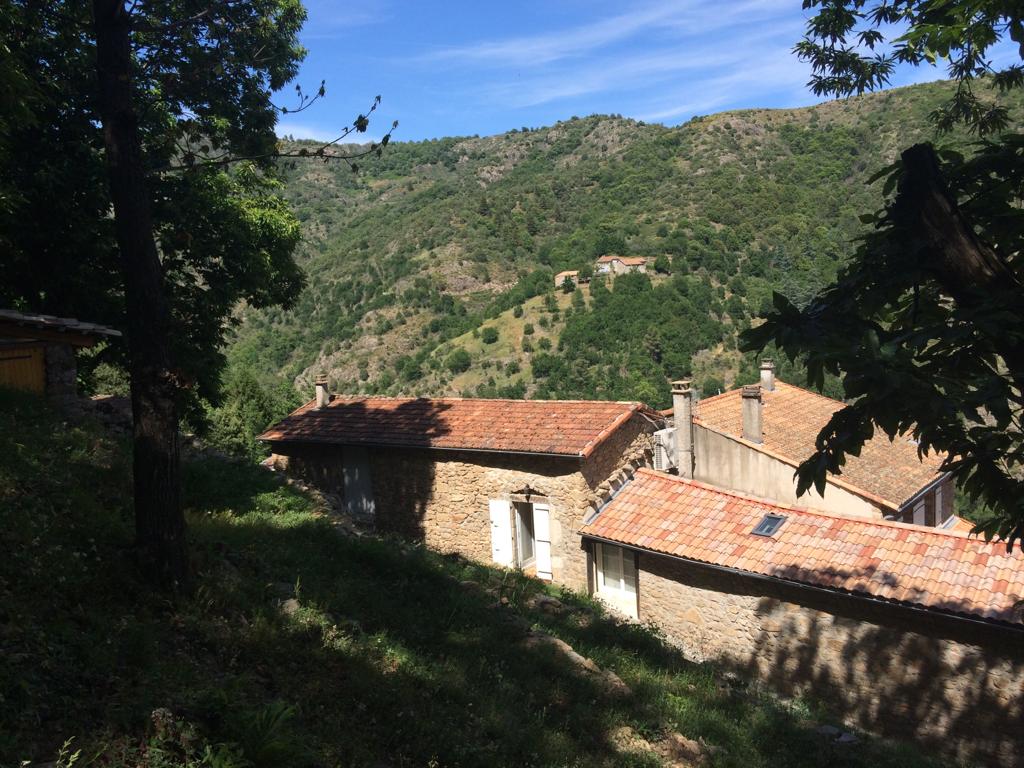 The image displays a scenic view of a stone building with a terracotta roof, nestled in a green valley. Rolling hills covered with trees rise in the background, under a clear blue sky, while gentle sunlight creates dappled shadows on the ground.