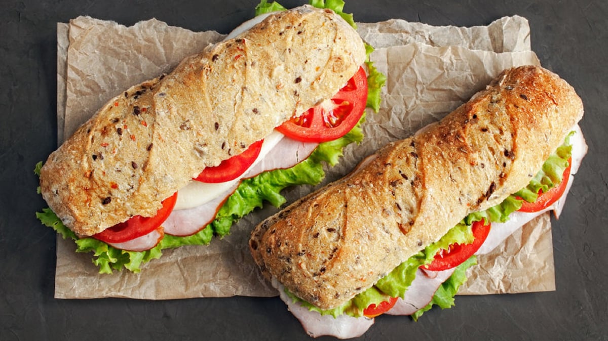 Two whole grain sandwiches are displayed on a piece of brown parchment paper. Each sandwich features layers of fresh lettuce, sliced tomatoes, and deli meats, all held together by a crusty, textured bread roll.