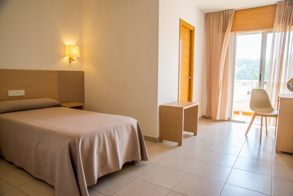 A simple room is presented with a single bed covered in a beige blanket. Light-colored walls complement the wooden furniture, including a bedside table and a chair. Natural light flows through the large window, framed by soft curtains, illuminating the space.