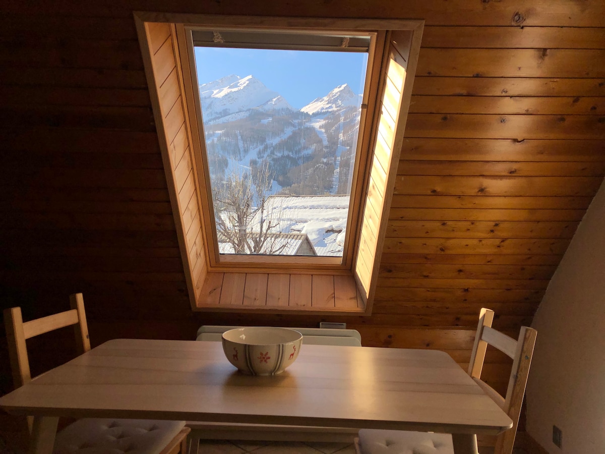 A wooden-paneled dining area features a simple table, complemented by two chairs. A large skylight offers a clear view of the mountains, allowing natural light to fill the space, while a bowl sits centrally on the table.