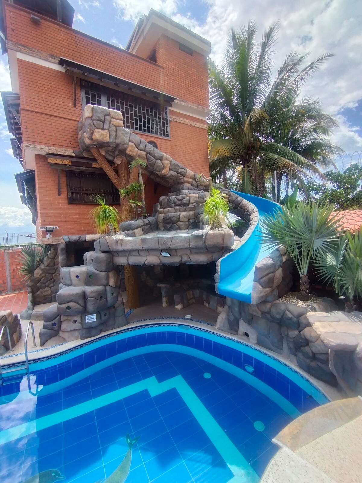 A curving water slide is seen cascading down a stone structure beside a blue swimming pool. Lush green palms and other plants frame the scene, contributing to the outdoor ambiance. A brick building stands in the background beneath a partly cloudy sky.