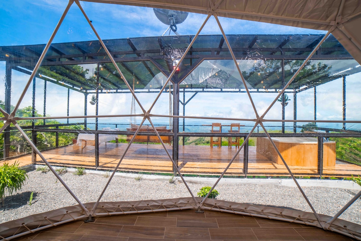 A view from inside the dome showcases a large outdoor terrace with wooden flooring and a dining table. The space is framed by geometric windows, allowing natural light to fill the interior. Lush greenery and distant hills are visible in the background.