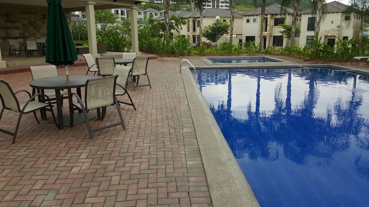 A patio area is shown, featuring a circular table with four chairs under a green umbrella. A swimming pool is visible beside the patio, reflecting the surrounding landscape and palm trees. The area is surrounded by well-maintained gardens and residential buildings in the background.