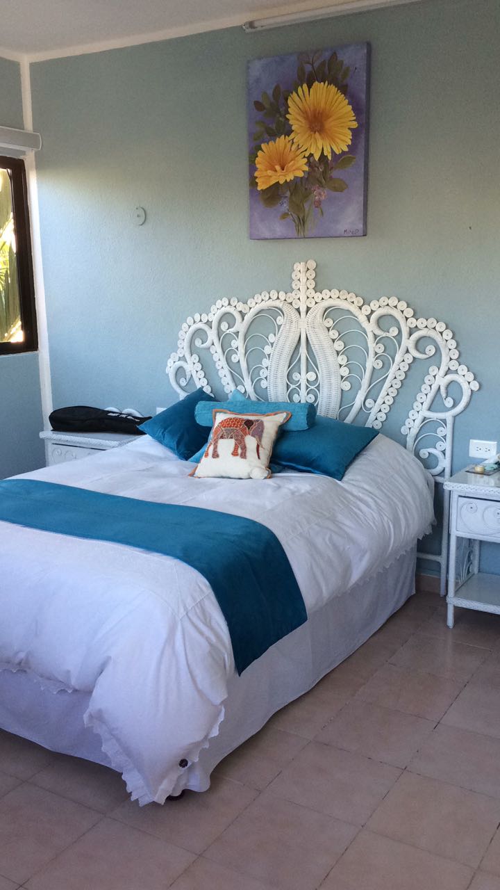 A serene bedroom features a white bed adorned with a teal accent blanket and a decorative pillow. A metal headboard adds an intricate touch. Natural light enters through a window adjacent to a floral artwork on the wall, creating a calm and inviting space.
