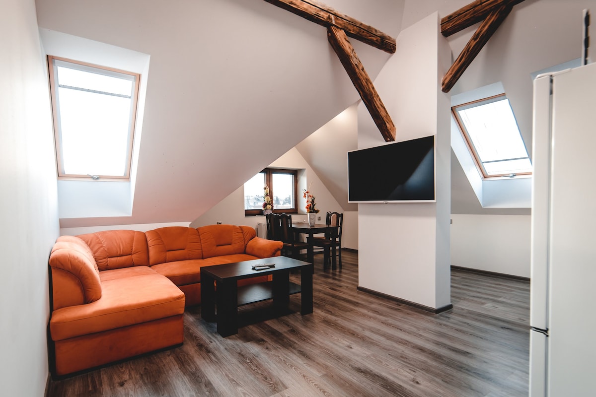 A modern living area is seen with a stylish orange sofa and a dark coffee table. Large windows allow natural light to fill the space, while exposed wooden beams add character. A dining table is positioned nearby, providing a comfortable setting for meals.