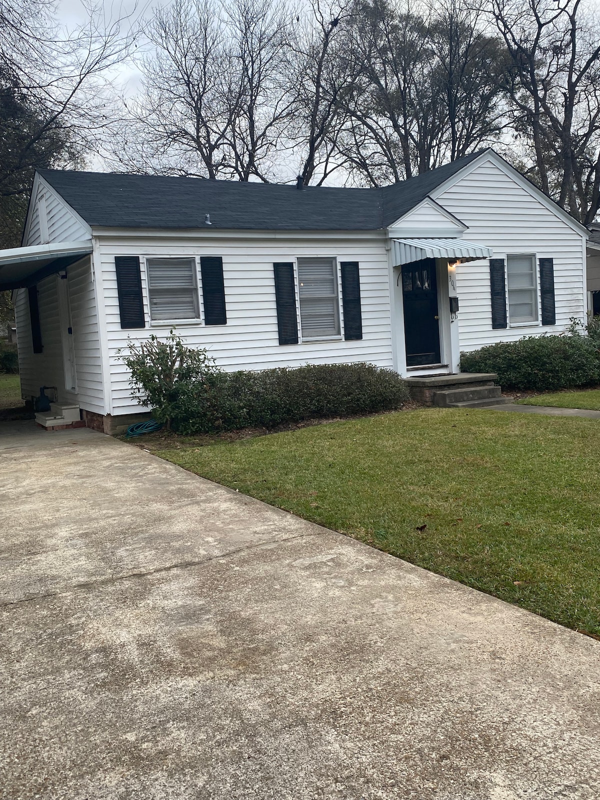 A cozy cottage-style house is presented, featuring white siding and a dark roof. Black shutters frame the windows, and the entrance is marked by a small porch. A landscaped front yard with green grass surrounds the house, alongside a concrete driveway leading to the carport.