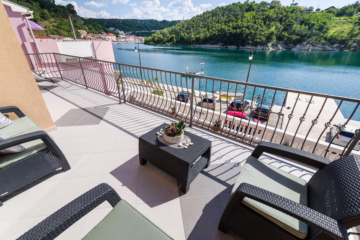 A spacious balcony area features wicker seating arrangements facing a tranquil water view. Decorative plants are arranged on a low table, and the nearby shoreline is visible across the calm water, with greenery lining the opposite bank.