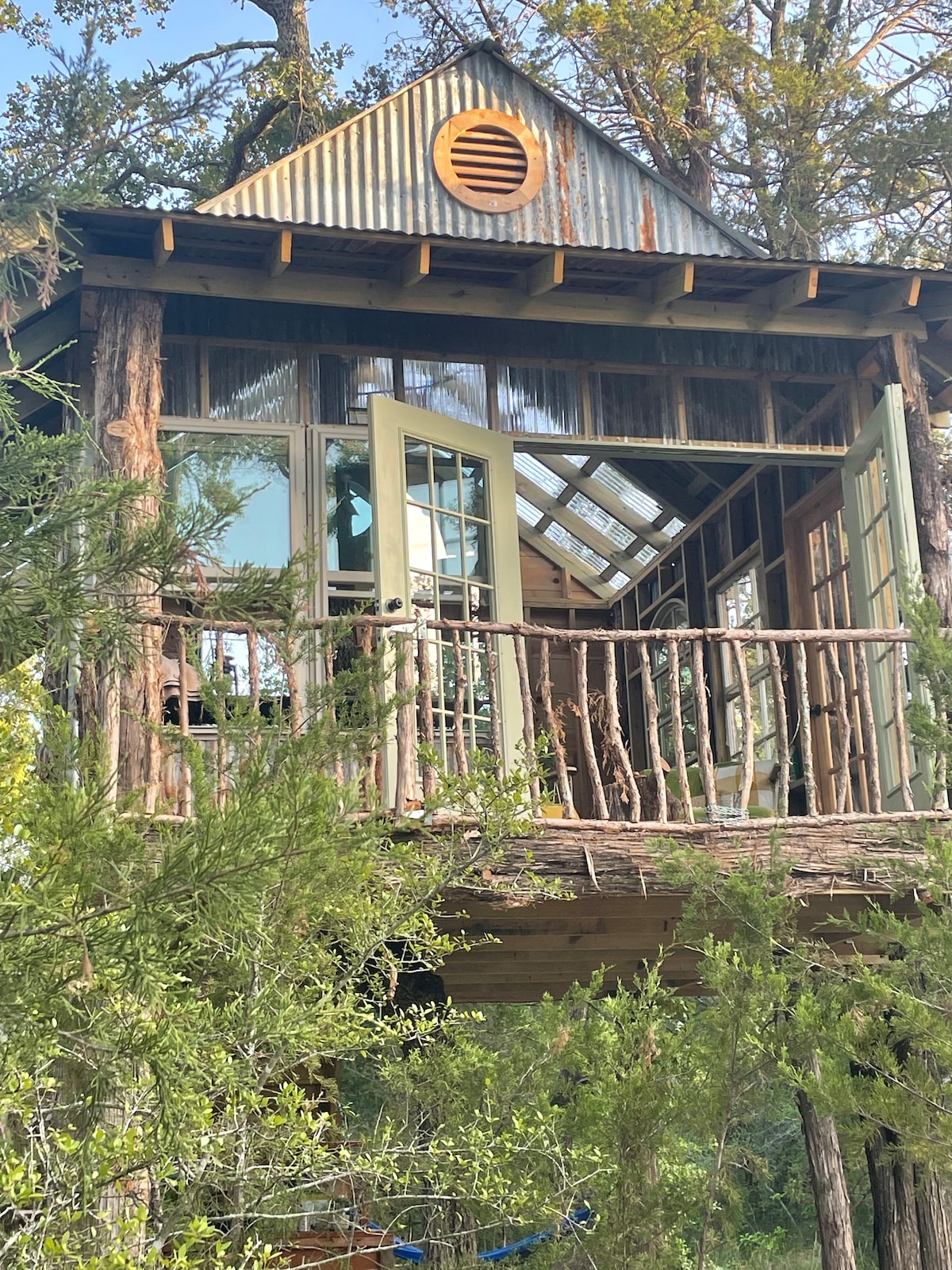 A treehouse elevated among trees features large glass doors opening onto a wooden balcony. Natural wood and rustic elements create a harmonious blend with the surroundings. The structure is surrounded by lush greenery, providing a secluded, serene escape.
